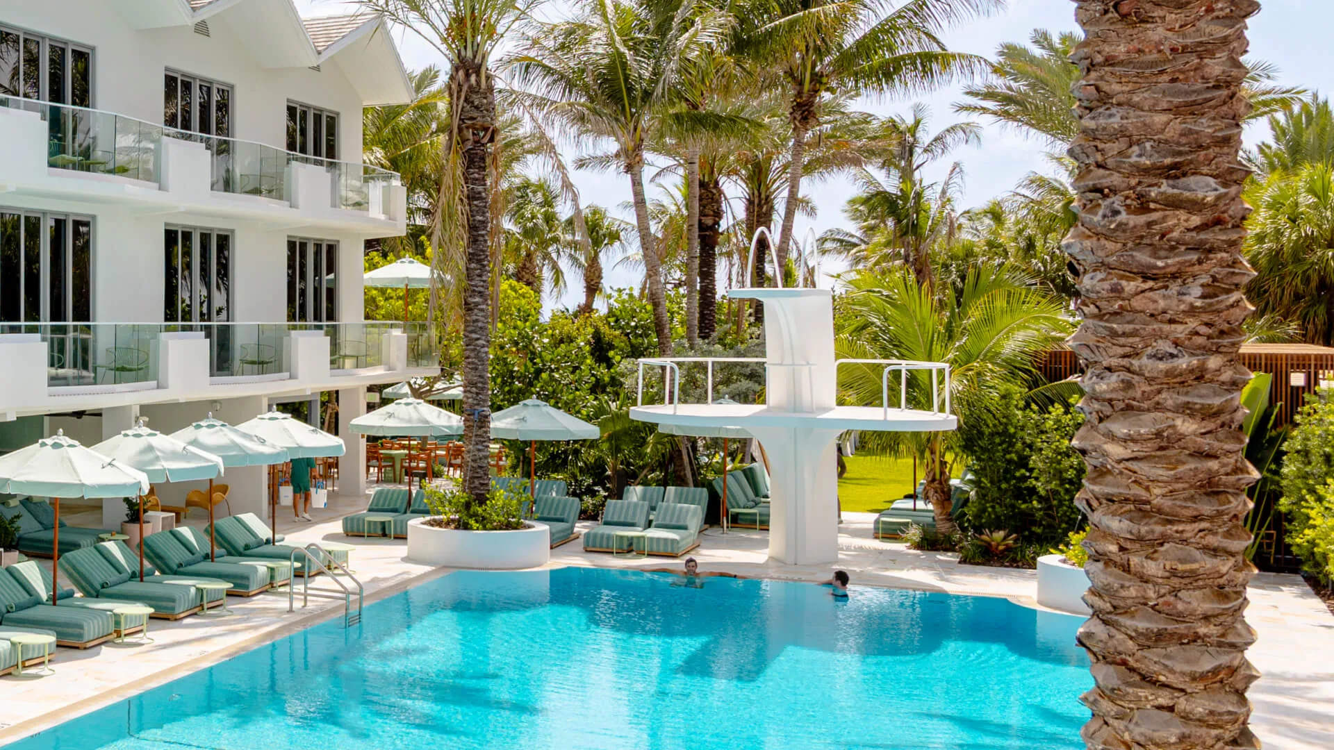 A pool with lounge chairs and umbrellas in front of a white building with balconies.