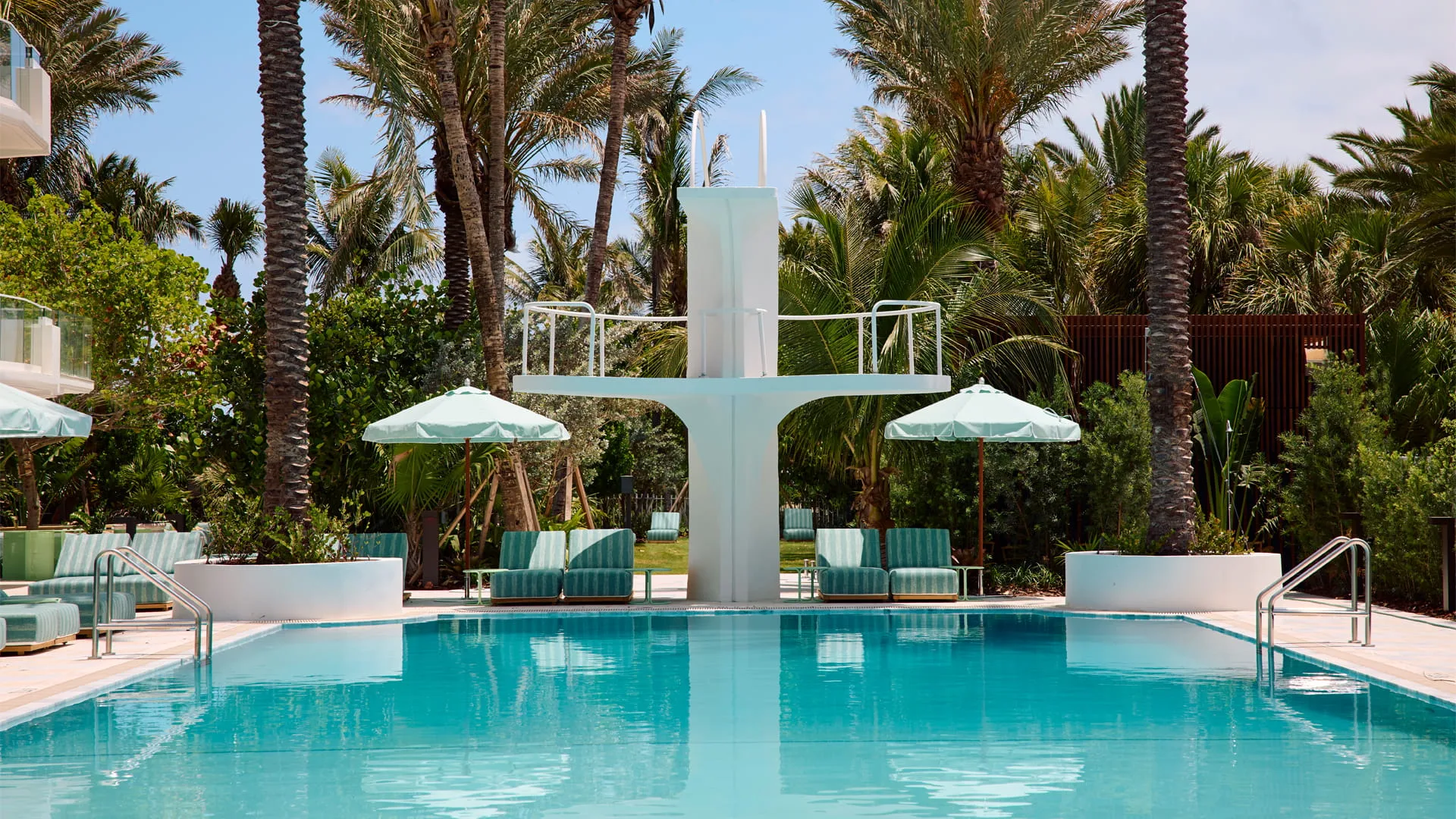 The Shelborne pool deck, turquoise water in front of the hotel's white Art Deco diving tower, with striped loungers and pale green parasols under tall palms.