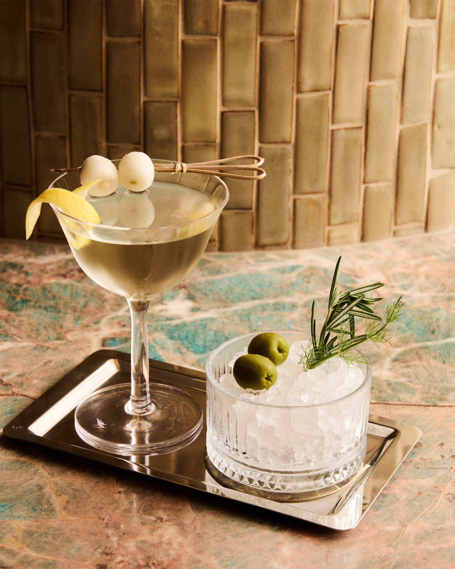 Martini cocktail with lemon twist and olives served alongside a chilled garnish glass on a marble bar top with warm textured tile background