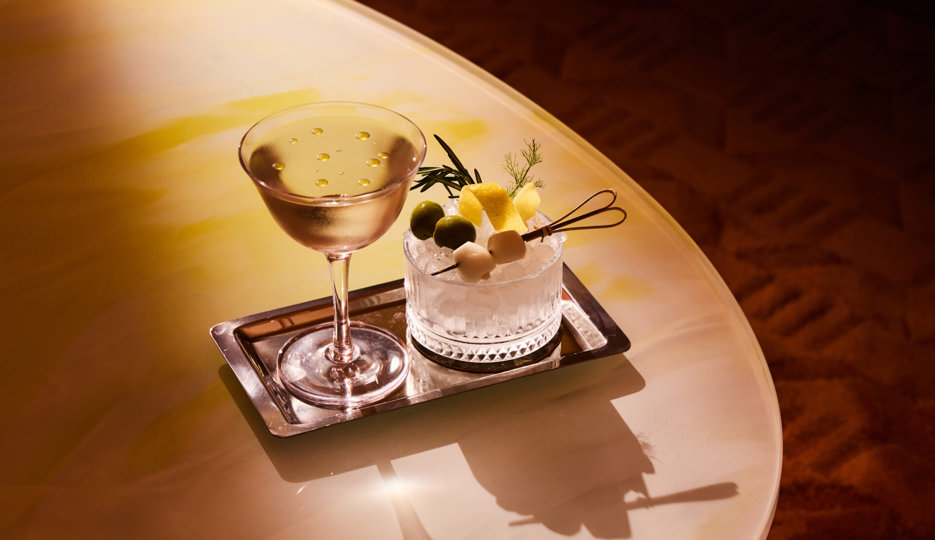 Elegant cocktails on a tray at The Shelborne bar with olives, citrus garnish, and warm ambient lighting